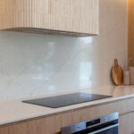 5144 Rossa Nova-Kristy Bunker project - Minimalist kitchen with a cooktop, wooden cabinetry, marble backsplash, and decorative cutting boards.