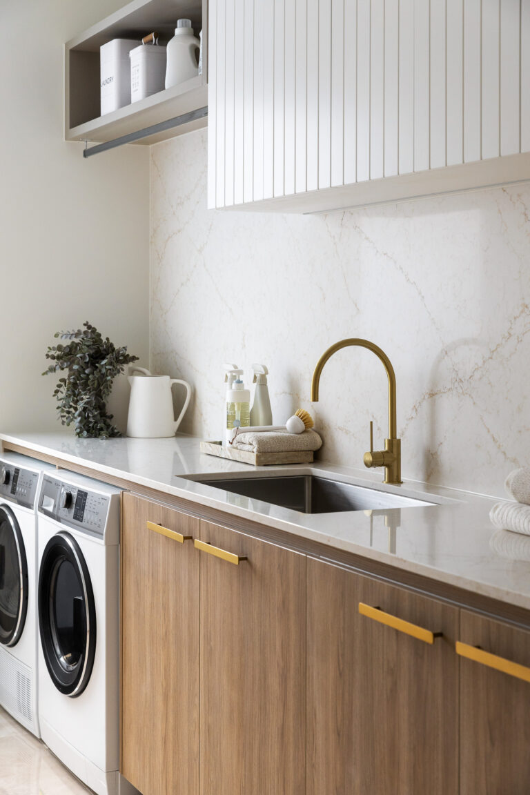 Caesarstone 5144 Rossa Nova surfaces in Modern laundry room with washer, dryer, wooden cabinets, marble countertop, gold faucet, and glass door.