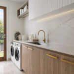 Caesarstone 5144 Rossa Nova surfaces in Modern laundry room with washer, dryer, wooden cabinets, marble countertop, gold faucet, and glass door.