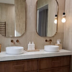Caesarstone 5144 Rossa Nova - Campaspe Kitchens Modern bathroom with double sinks, two mirrors, wooden vanity, hanging lights, and beige tiles.