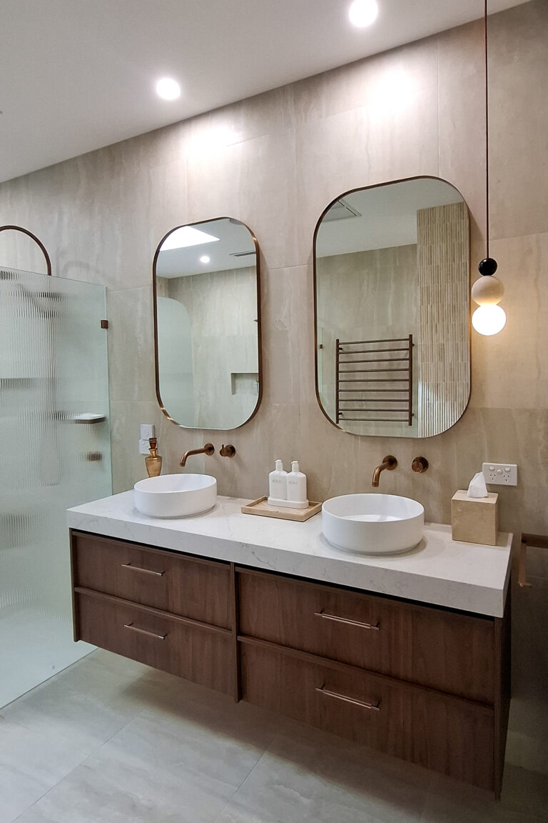 Caesarstone 5144 Rossa Nova - Campaspe Modern bathroom with double sinks, round mirrors, wooden cabinets, pendant light, and glass shower enclosure.