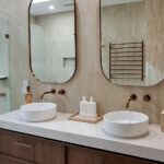 Caesarstone 5144 Rossa Nova - Campaspe Modern bathroom with double sinks, round mirrors, wooden cabinets, pendant light, and glass shower enclosure.